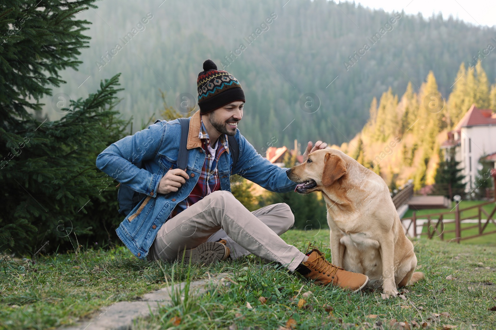 Photo of Happy man and adorable dog sitting on green grass in mountains. Traveling with pet