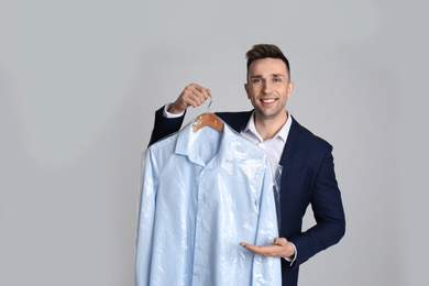 Photo of Man holding hanger with shirt in plastic bag on light grey background. Dry-cleaning service