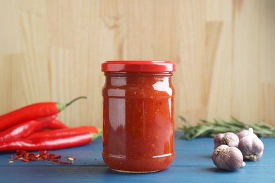 Spicy chili sauce in jar, garlic and peppers on blue wooden table