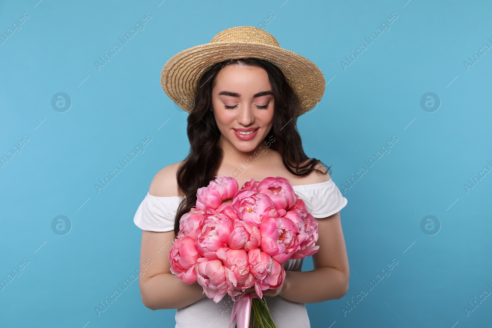 Photo of Beautiful young woman in straw hat with bouquet of pink peonies against light blue background