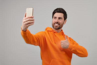 Photo of Smiling man taking selfie with smartphone and showing thumbs up on grey background