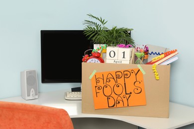 Photo of Box with different items and words Happy Fool's Day at workplace in office