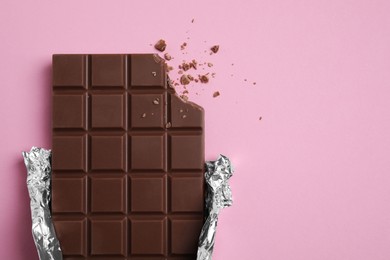 Photo of Bitten milk chocolate bar wrapped in foil on pink background, top view. Space for text