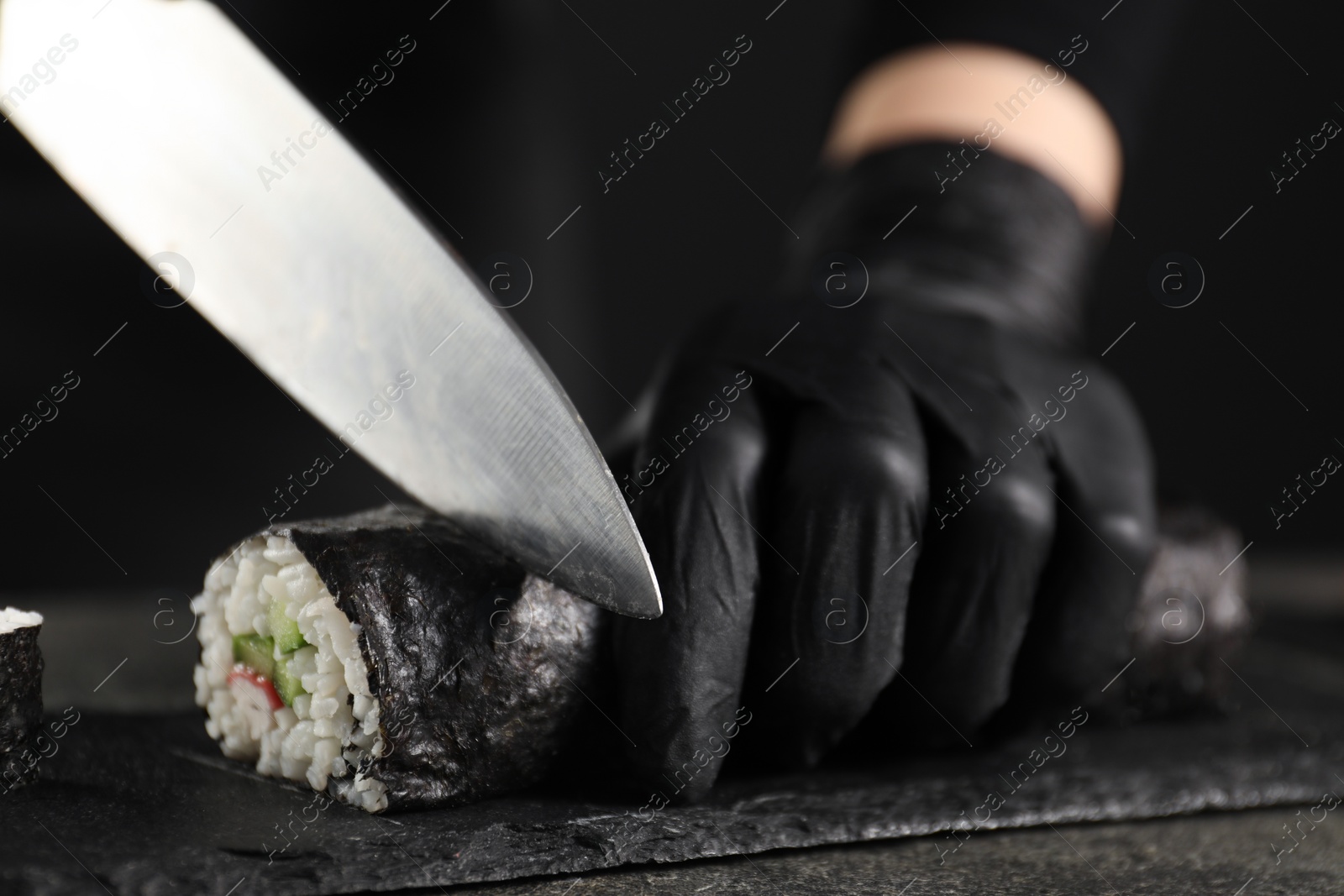 Photo of Chef in gloves cutting sushi roll at dark table, closeup