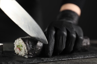 Photo of Chef in gloves cutting sushi roll at dark table, closeup