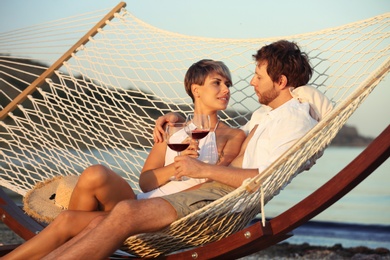 Photo of Young couple resting with glasses of wine in hammock on beach
