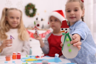 Photo of Cute little children with beautiful Christmas crafts at table in room, focus on paper snowman. Space for text