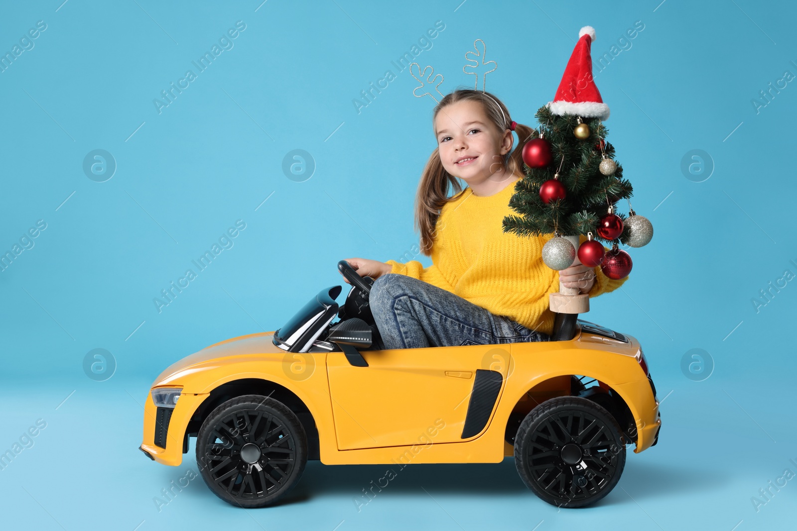 Photo of Cute little girl with Christmas tree driving children's electric toy car on light blue background