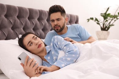Photo of Young woman ignoring her distrustful boyfriend while using smartphone in bed at home