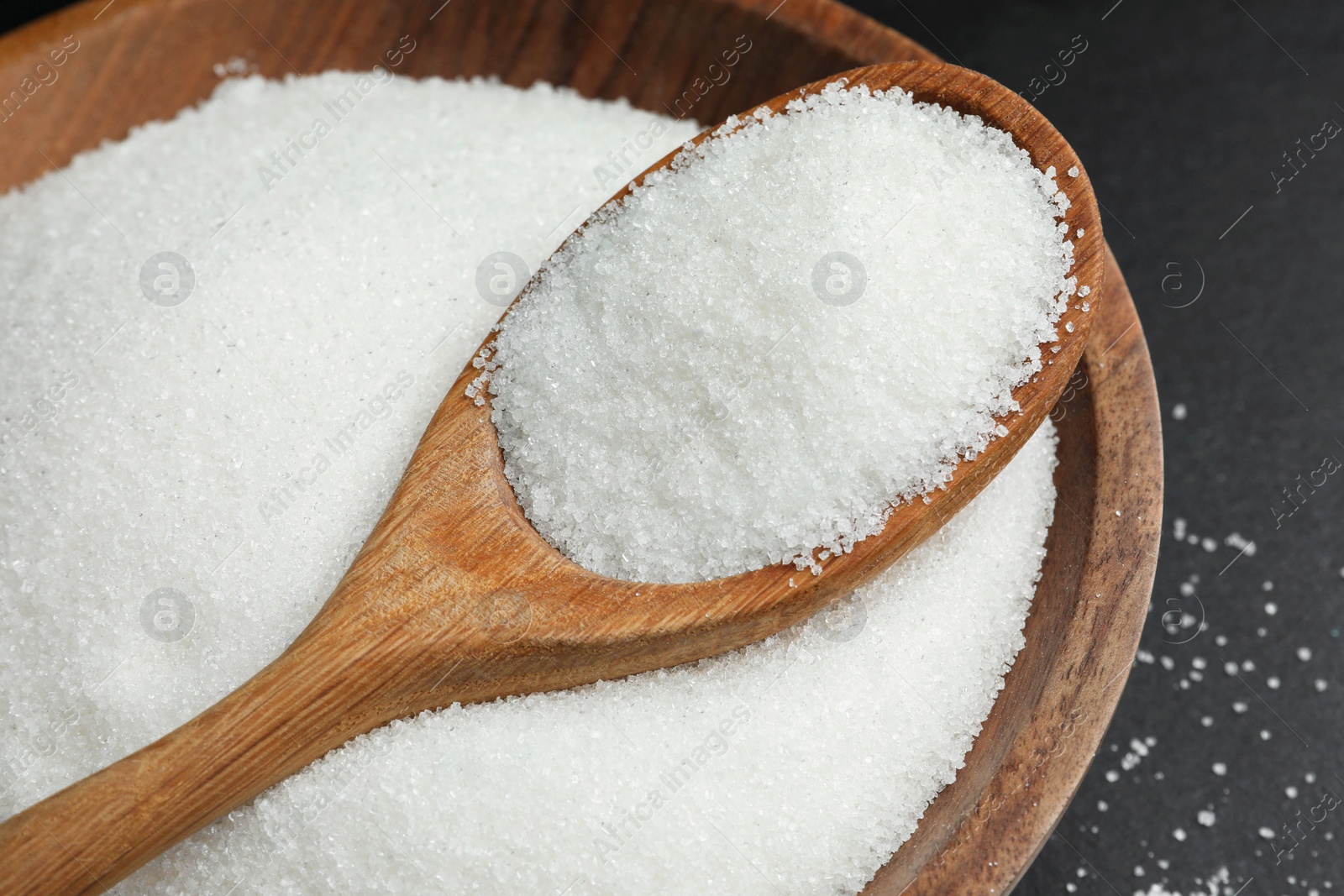 Photo of Granulated sugar in bowl and spoon on black table, closeup