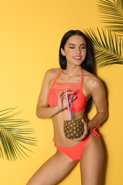Photo of Beautiful young woman in stylish bikini with cocktail and tropical leaves on yellow background
