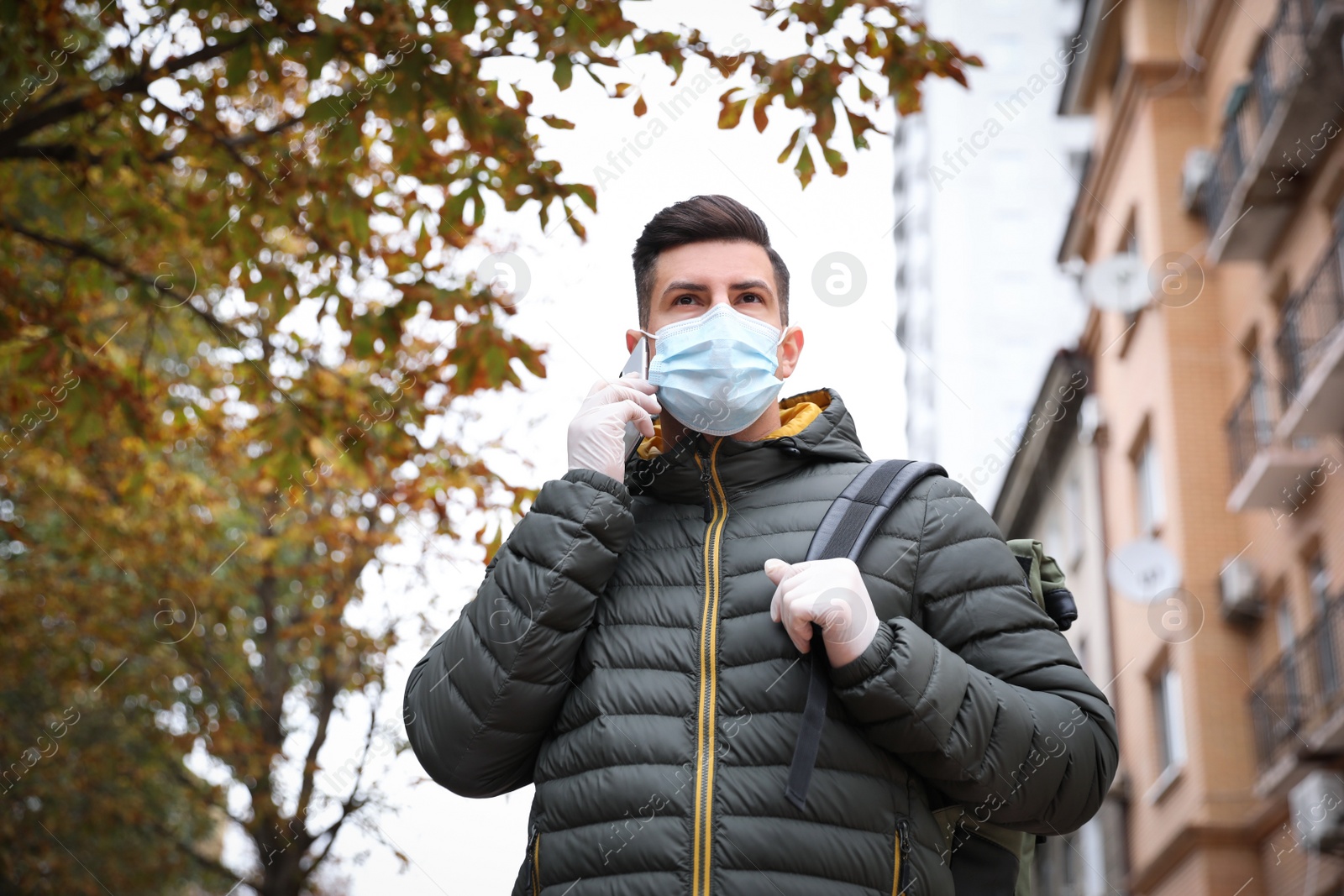 Photo of Man in medical face mask and gloves talking on phone while walking outdoors. Personal protection during COVID-19 pandemic