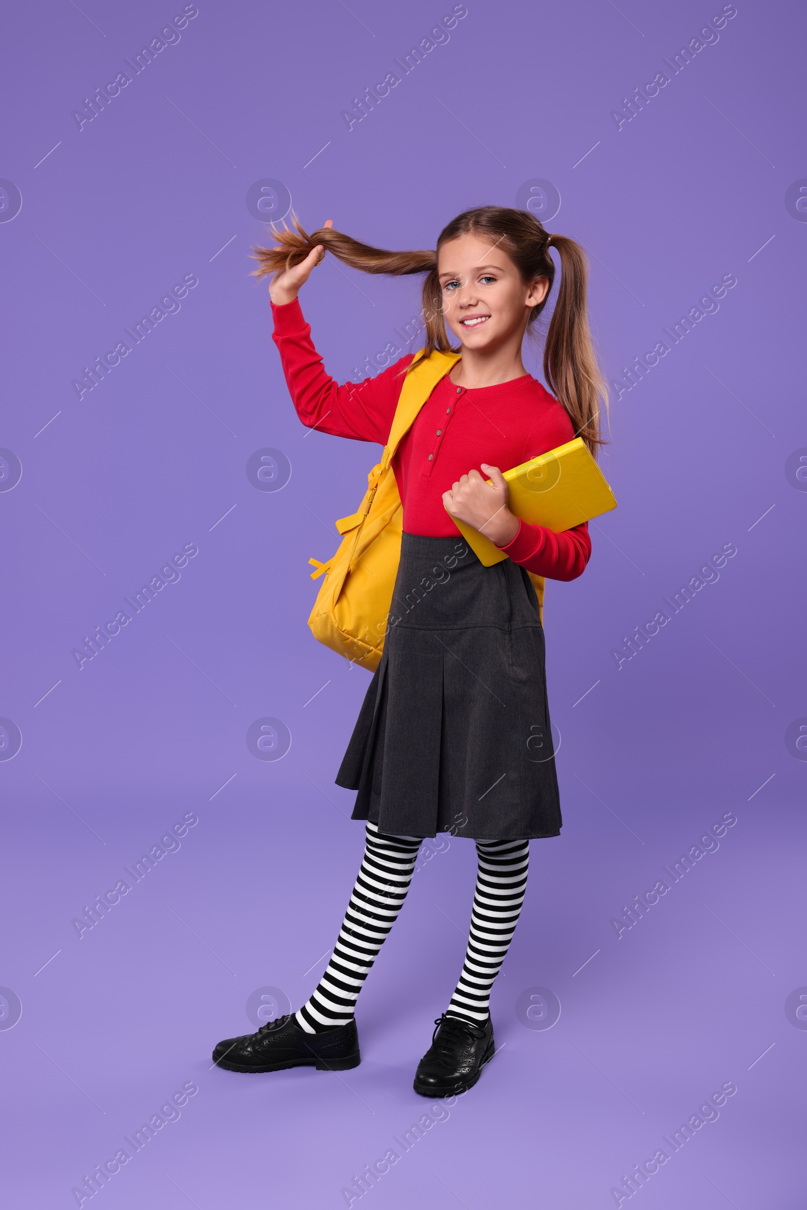 Photo of Smiling schoolgirl with backpack and book on violet background