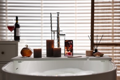 White wooden tray with smartphone, burning candles and beauty products on bathtub in bathroom