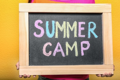 Photo of Child holding blackboard with words SUMMER CAMP chalked in different colors, closeup