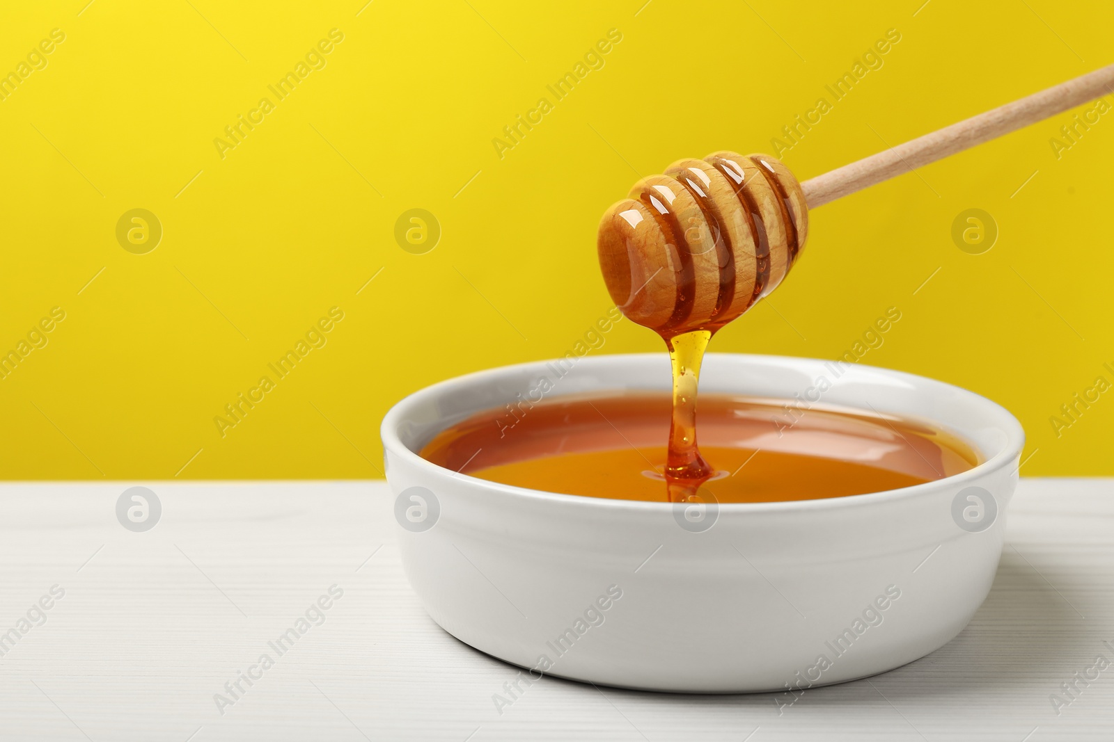 Photo of Pouring tasty honey from dipper into bowl at white wooden table against yellow background, closeup. Space for text