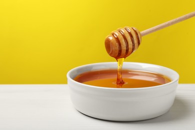 Pouring tasty honey from dipper into bowl at white wooden table against yellow background, closeup. Space for text