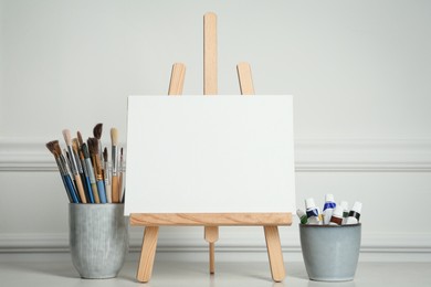 Photo of Easel with blank canvas, paints and brushes on white wooden table