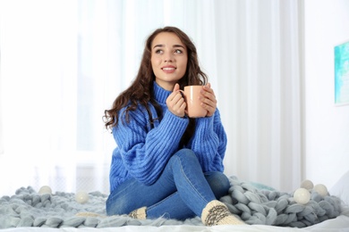 Photo of Young beautiful woman in warm sweater with cup of hot drink on bed at home