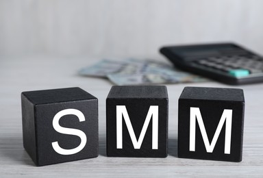 Photo of Black cubes with abbreviation SMM (Social media marketing), money and calculator on white wooden table