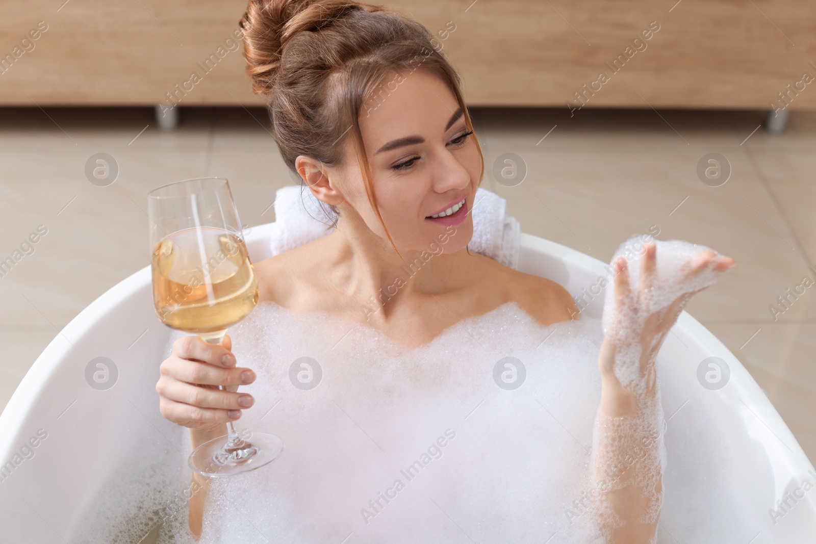 Photo of Beautiful woman with glass of wine enjoying bubble bath at home