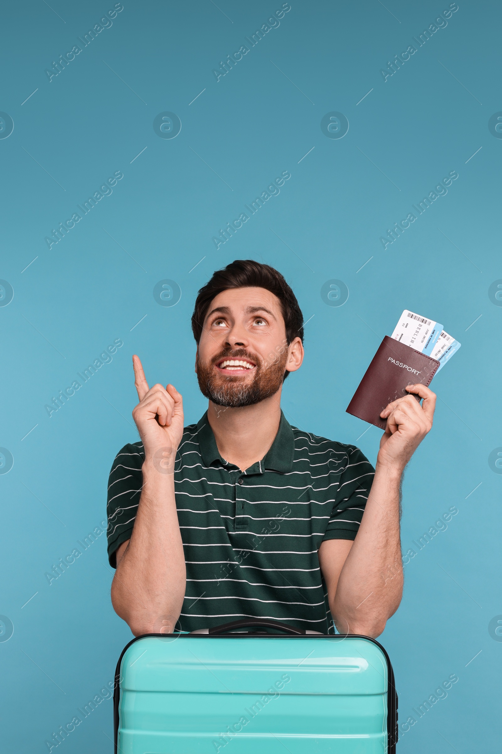 Photo of Smiling man with passport, suitcase and tickets pointing at something on light blue background. Space for text