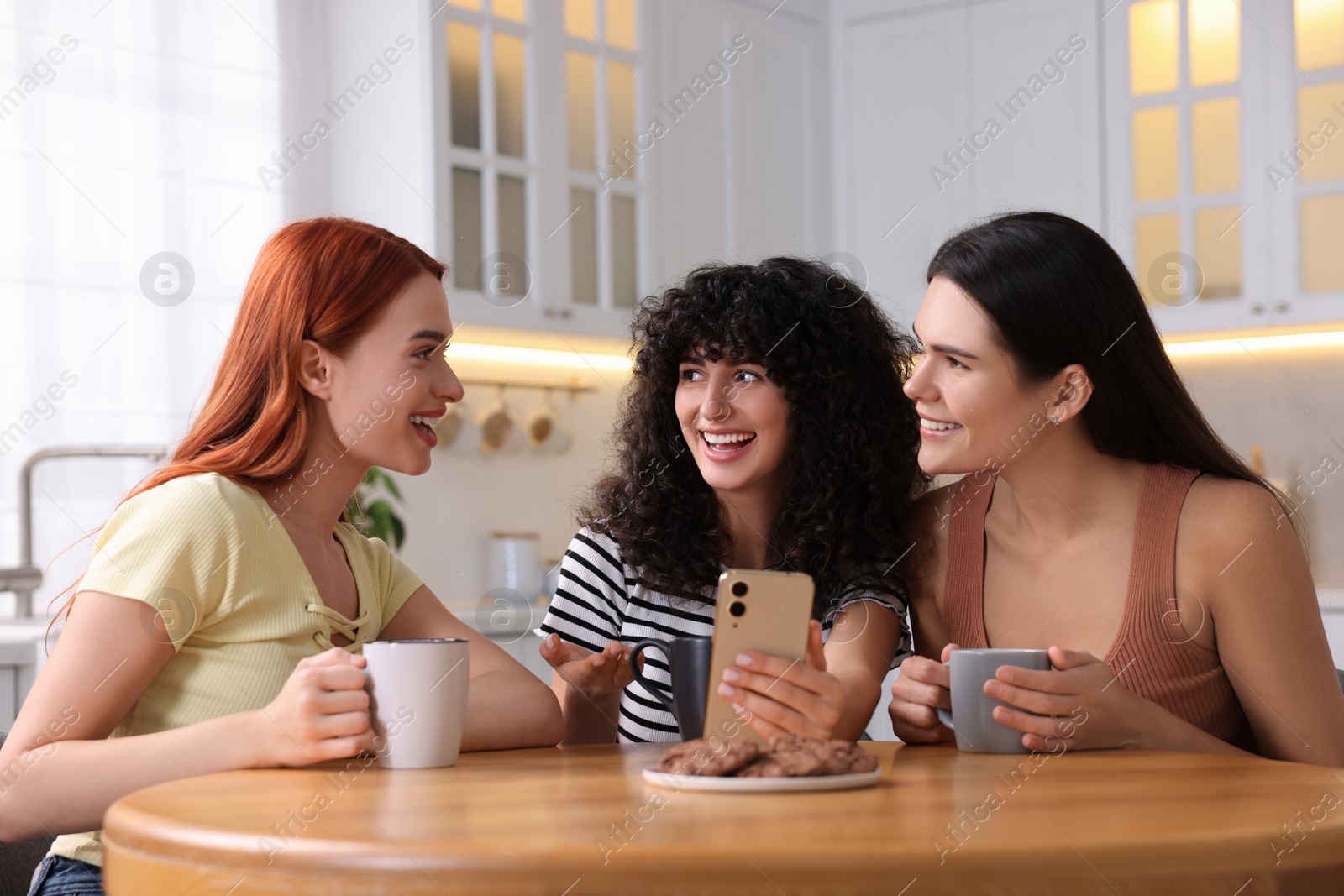 Photo of Happy young friends with smartphone spending time together at table in kitchen