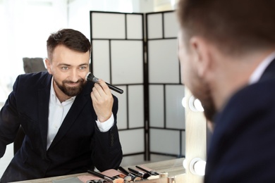 Photo of Young man using makeup brush near mirror in dressing room