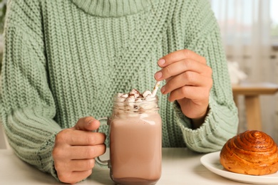 Woman holding mason jar of delicious cocoa drink with marshmallows at white table, closeup