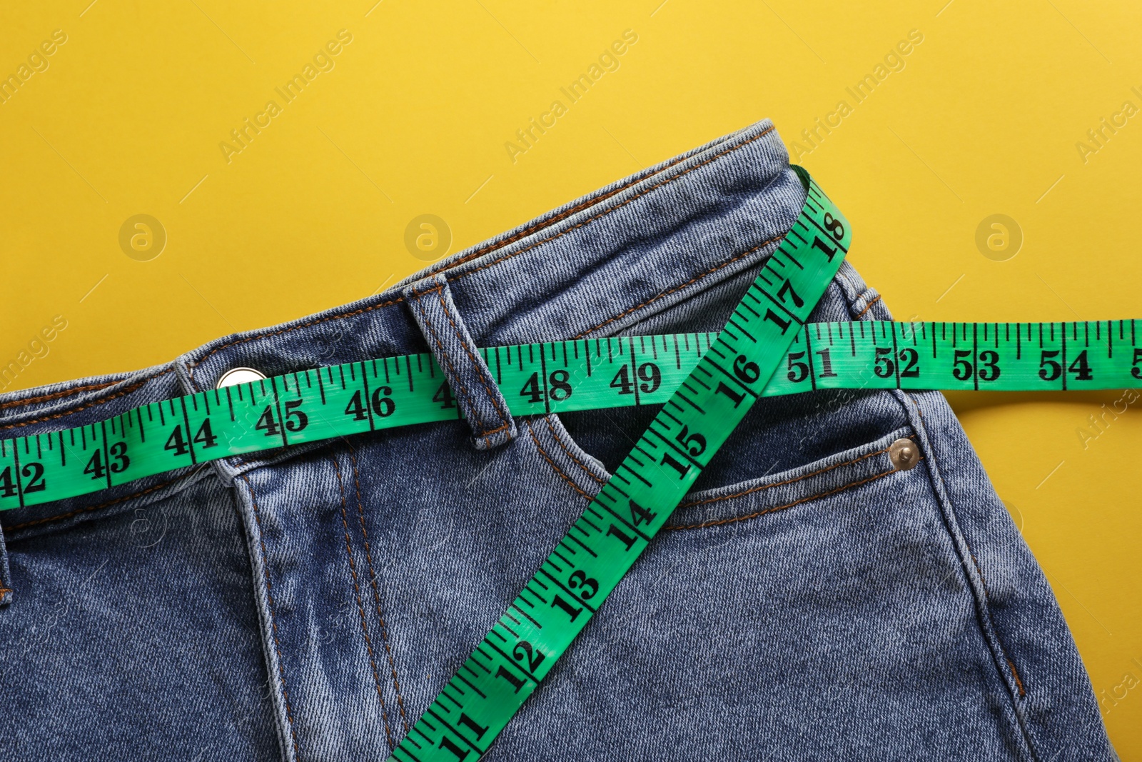 Photo of Jeans with measuring tape on yellow background, top view. Weight loss concept