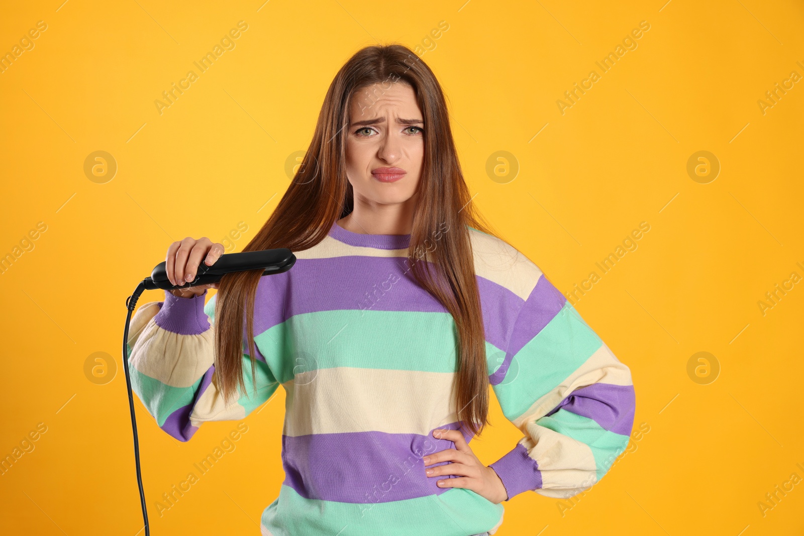 Photo of Upset young woman with flattening iron on yellow background. Hair damage