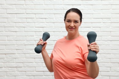 Photo of Happy overweight woman doing exercise with dumbbells near white brick wall, space for text