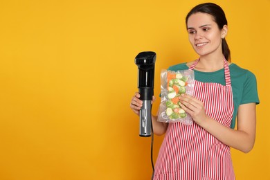 Photo of Beautiful young woman holding sous vide cooker and vegetables in vacuum pack on orange background. Space for text