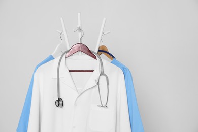 Photo of Medical uniforms and stethoscope hanging on rack against light grey background