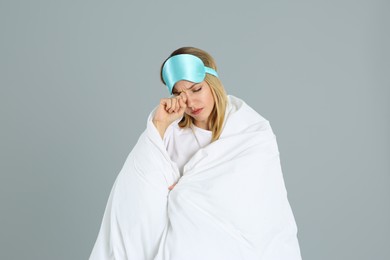Young woman in sleeping mask wrapped with soft blanket on grey background