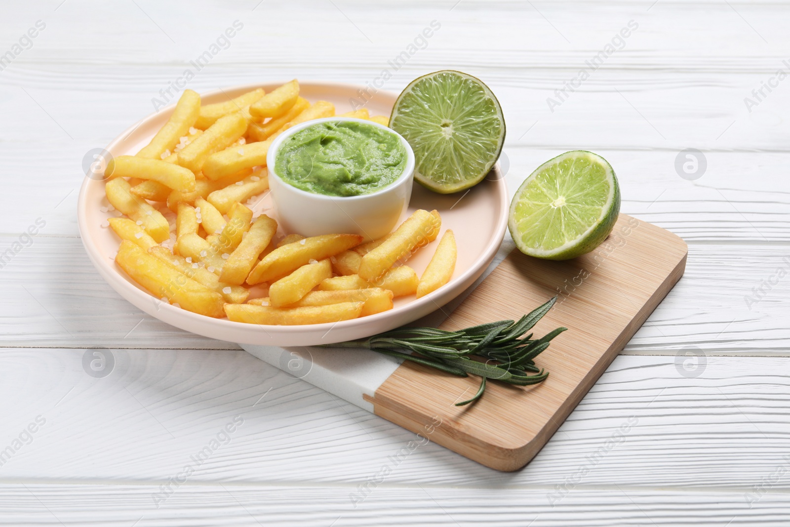 Photo of Tray with plate of french fries, avocado dip, lime and rosemary on white wooden table