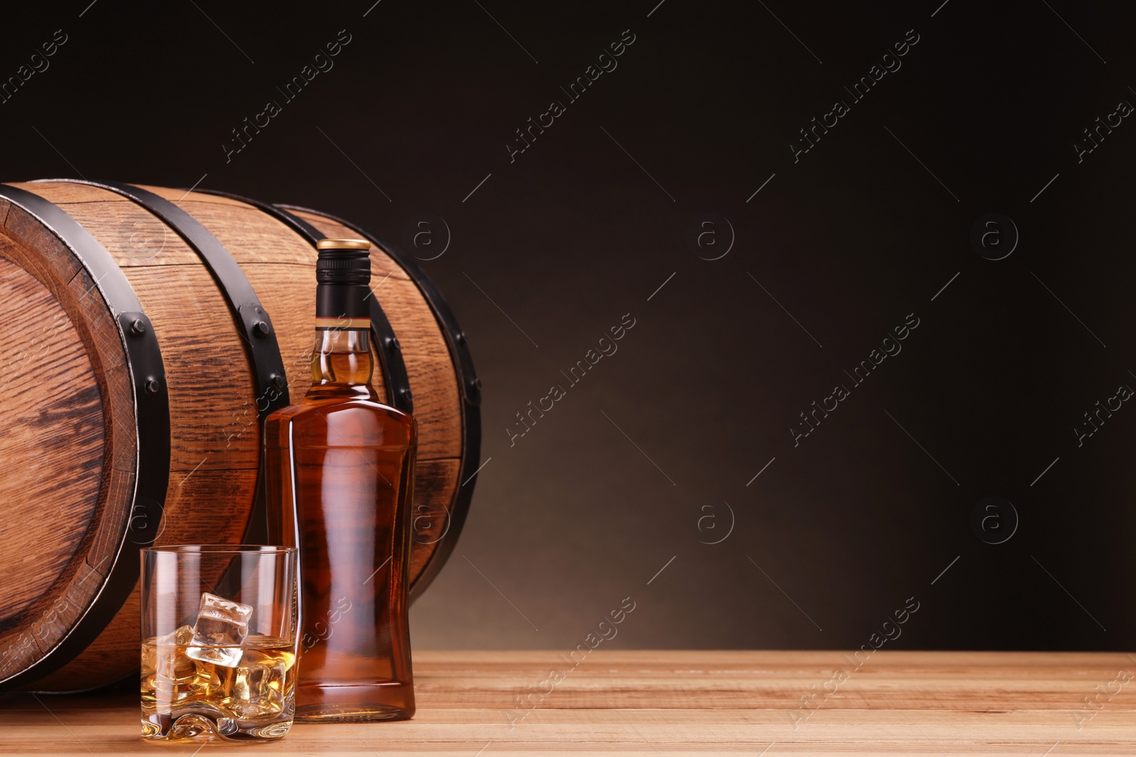 Photo of Whiskey with ice cubes in glass, bottle and barrel on wooden table against dark background, space for text