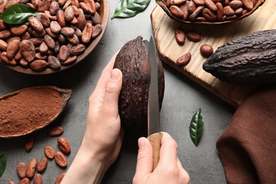 Woman cutting ripe cocoa pod over grey table with products, top view