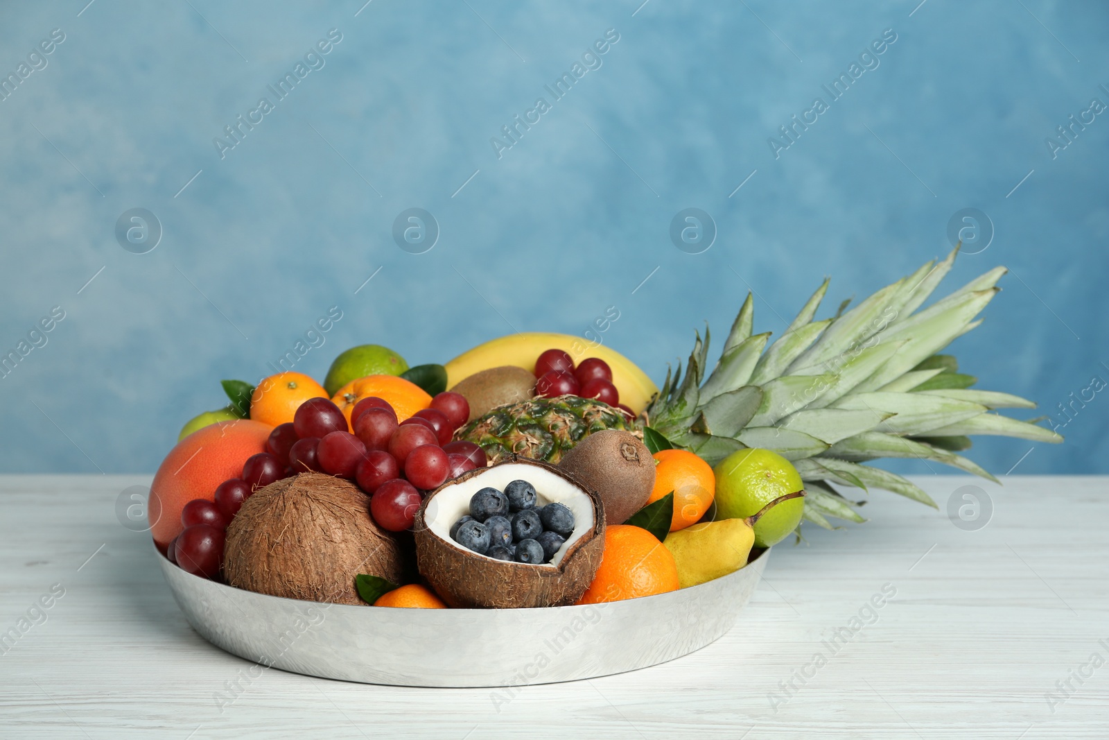 Photo of Assortment of fresh exotic fruits in metal tray on white wooden table against light blue background