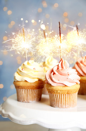 Delicious birthday cupcakes with sparklers on stand against blurred background