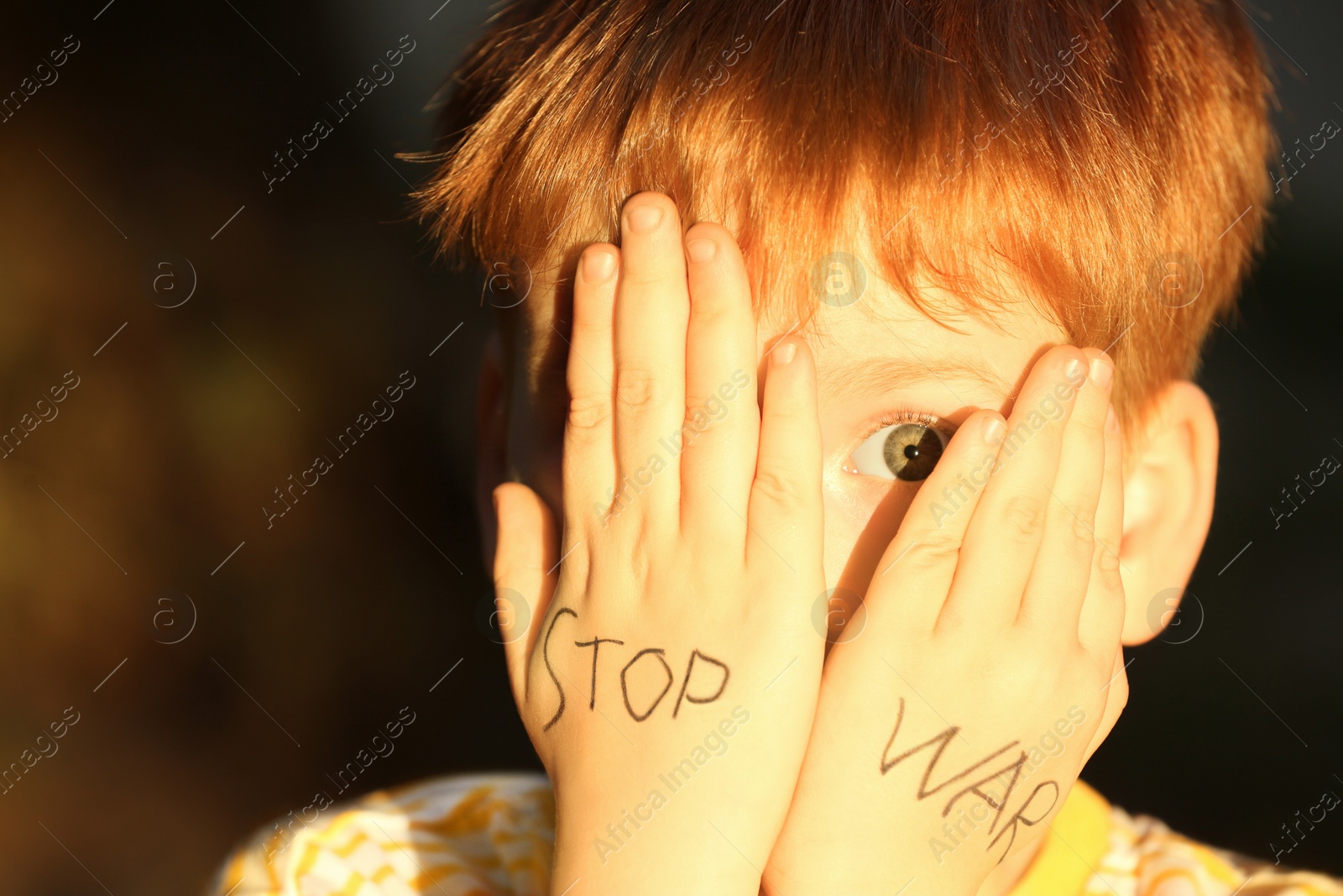 Photo of Little boy hiding face and words Stop War written on his hands outdoors, closeup
