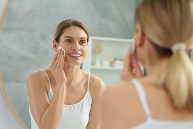 Photo of Beautiful woman removing makeup with cotton pad near mirror indoors
