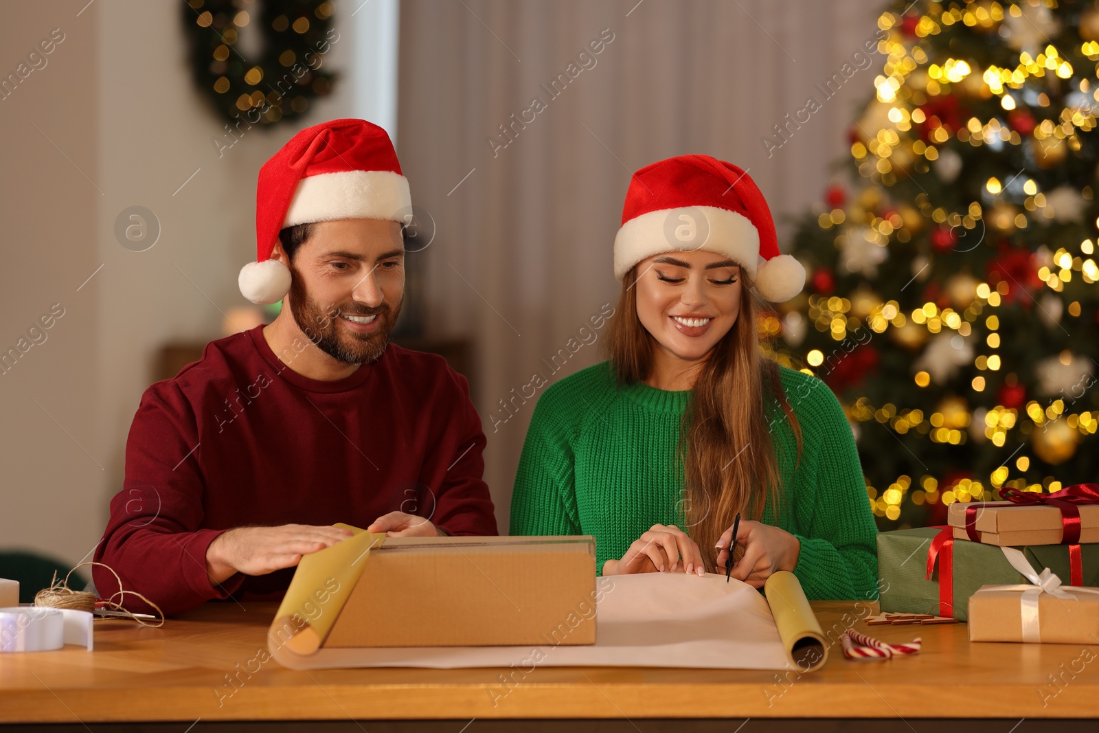 Photo of Happy couple in Santa hats decorating Christmas gift with wrapping paper at table in room