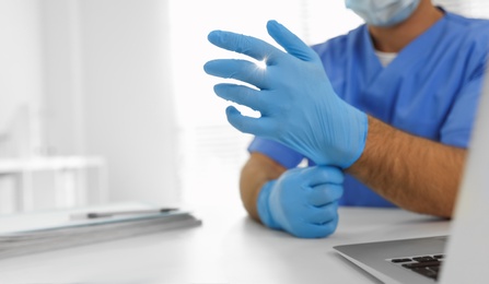 Photo of Doctor in protective mask putting on medical gloves at table in office, closeup. Space for text