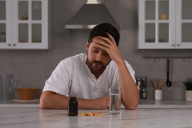 Depressed man with glass of water and antidepressant pills at table in kitchen