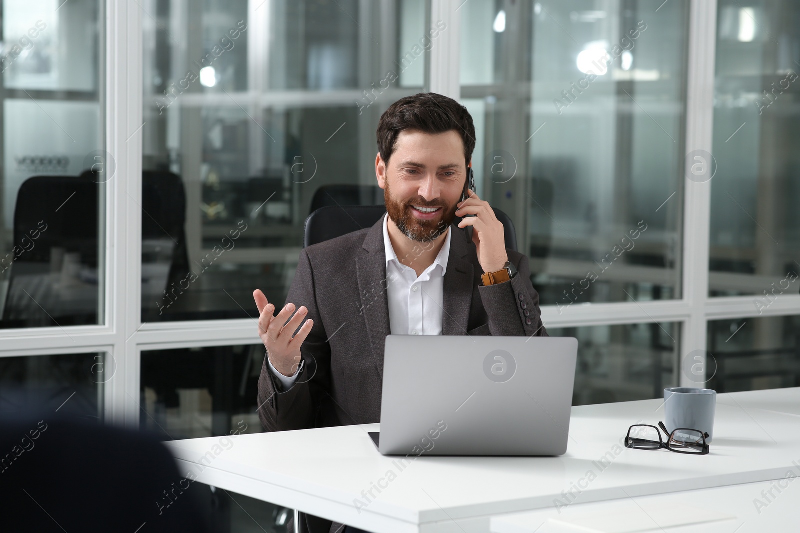 Photo of Man talking on phone while working with laptop at white desk in office