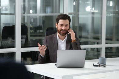 Photo of Man talking on phone while working with laptop at white desk in office