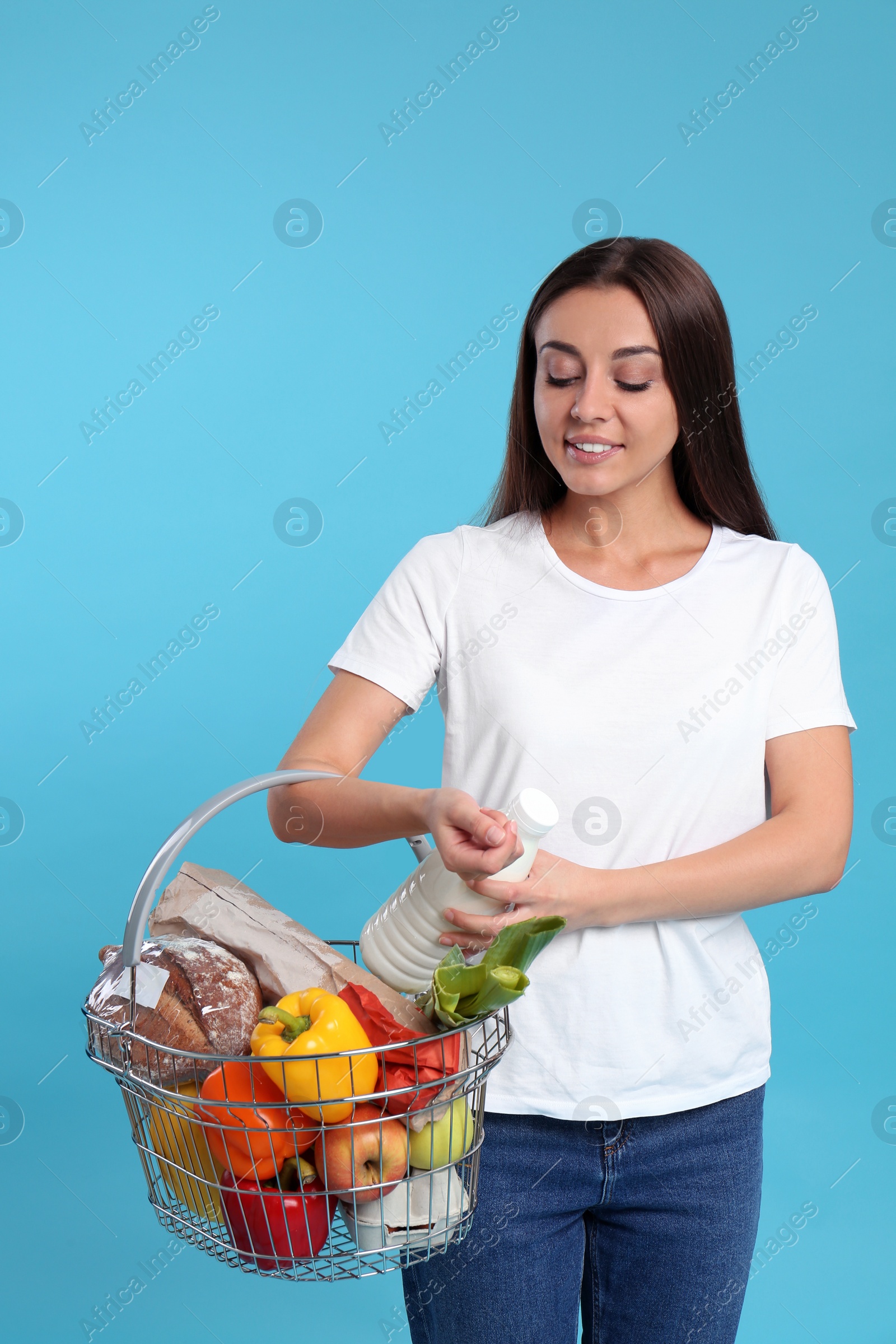 Photo of Young woman with shopping basket full of products on blue background