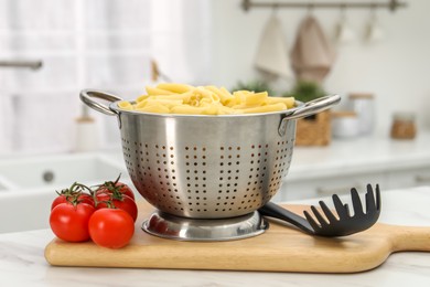 Cooked pasta in metal colander and tomatoes on light marble table in kitchen, closeup
