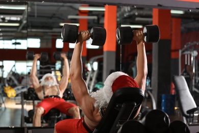 Photo of Young muscular man in Santa costume training with dumbbells at modern gym